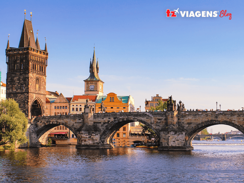 Um dos pontos turísticos de Praga que devem ser visitados é a Ponte Carlos (Charles Bridge). Na foto, a ponte e sua torre, num dia claro, com céu azul claro.