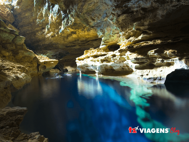Um bom lugar para se conhecer nas férias de Julho é Chapada Diamantina. Imagem de uma gruta com um lago dentro, a água do lago tem duas tonalidades de azul. 