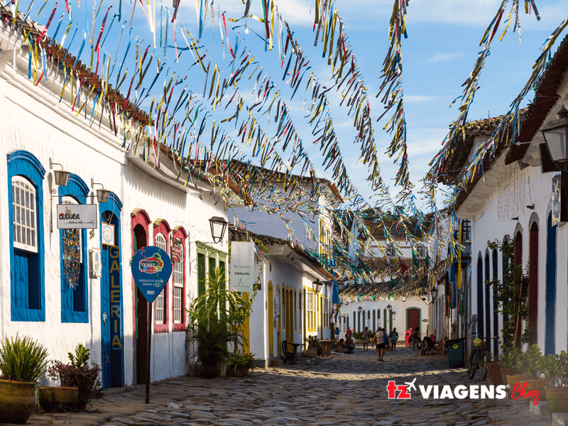 Nas férias de julho, você deve visitar Paraty. Na imagem, uma rua histórica de Paraty, casas brancas com portas e janelas coloridas e várias bandeirinhas penduradas. Pessoas ao fundo andando ou sentadas.