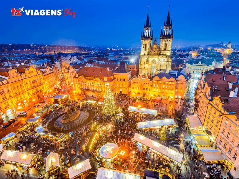 Entre os pontos turísticos de Praga, está a Oldtown Square. A imagem aérea noturna, a praça toda iluminada, enfeites de natal, várias barracas e com movimentação bem grande de pessoas. 