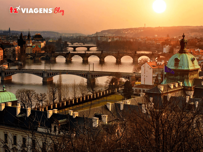 Lista de Pontos turísticos de Praga. Imagem aérea do rio Moldava, suas margens com casas e pontos turísticos. Três pontes em quadro e um lindo céu amarelo, com um por do sol.