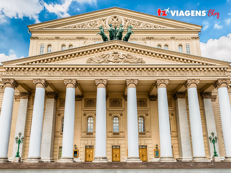 Foto da fachada do grande Teatro Bolshoi, em Moscou e aos fundo um céu azulado com algumas nuvens. O Teatro Bolshoi faz parte da lista de 10 Pontos turísticos da Rússia que você precisa conhecer.
