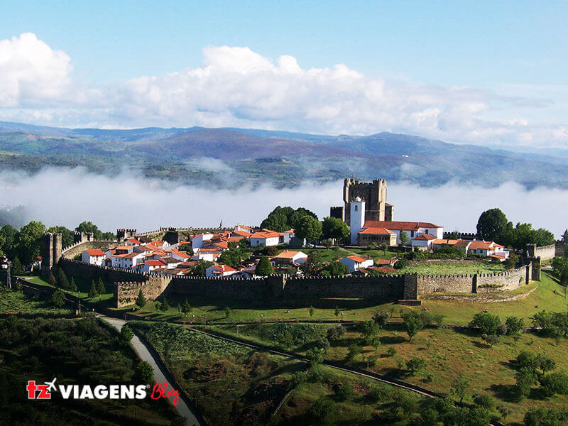 Bragança é um dos pontos do norte em uma Viagem para Portugal. Na imagem, uma vila cercada por uma muralha de um castelo, em um monte. Nuvens e céu azul em cima da vila.