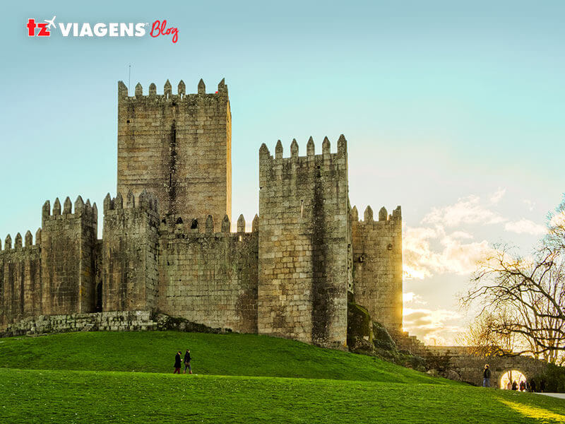 Guimaraes é uma cidade do norte de Portugal. É um ponto para conhecer em uma Viagem para Portugal. Na foto a imagem do castelo da cidade. Muralhas do castelo sob um gramado verde e o céu bem azulado.