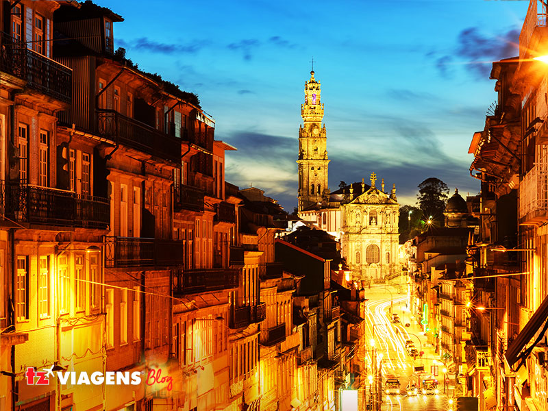 Torre dos Clérigos em Porto. , Portugal. É um ponto para conhecer em uma Viagem para Portugal. Na imagem prédios históricos, uma grande avenida e ao fundo a Torre dos Clérigos. Foto noturna, luzes acesas e o céu azul escuro.